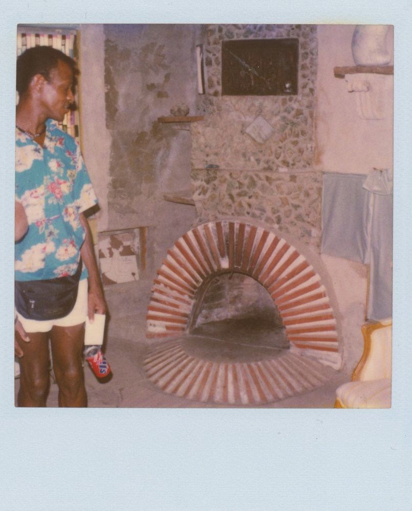 Download the full-sized image of A Photograph of Marsha P. Johnson Wearing a Hawaiian Shirt and Fanny Pack, Standing Near a Brick Fireplace