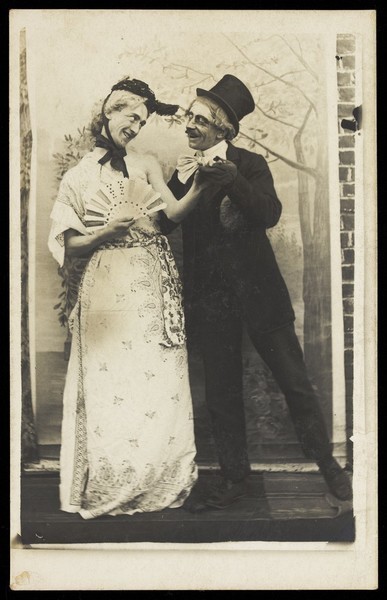 Download the full-sized image of Two amateur actors, one in drag as 'Charley's Aunt', greet each other in front of painted scenery. Photographic postcard, 191-.